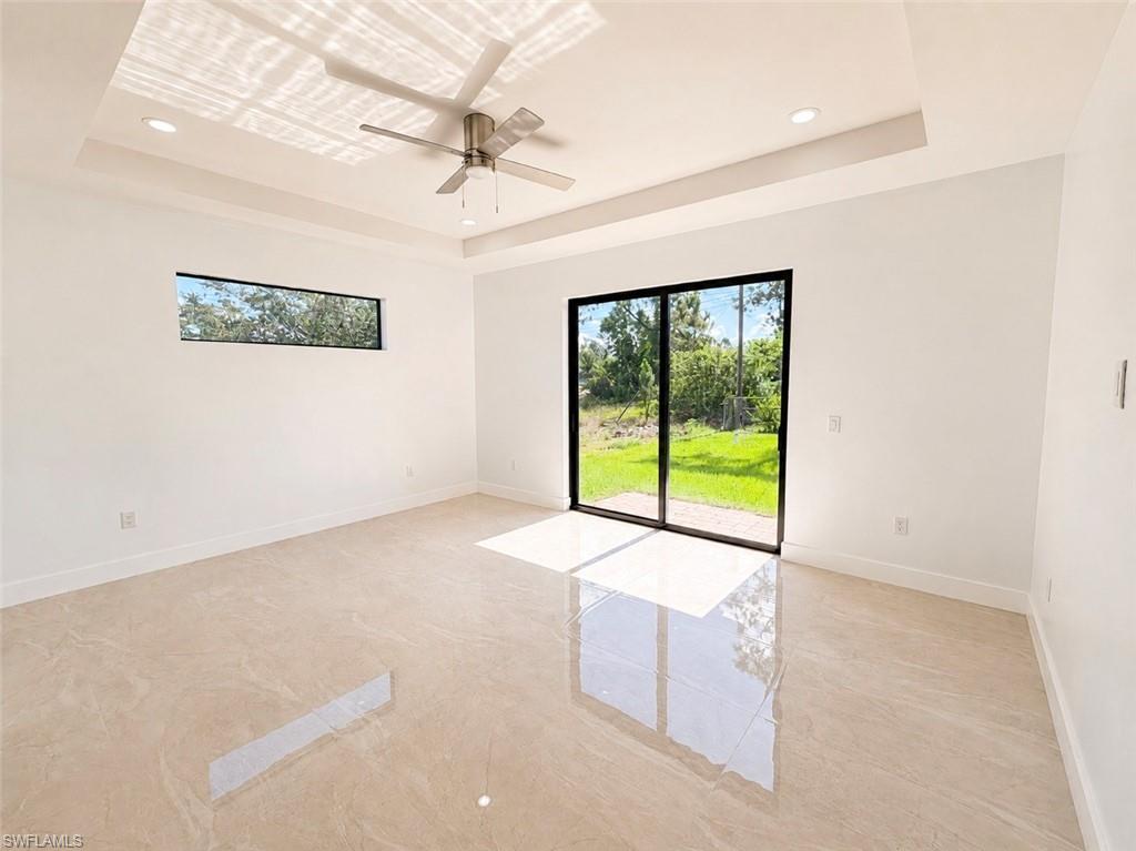 Spacious interior featuring a tray ceiling with recessed lighting, a modern ceiling fan, large format tile flooring, a horizontal window, and sliding glass doors opening to outdoor green space