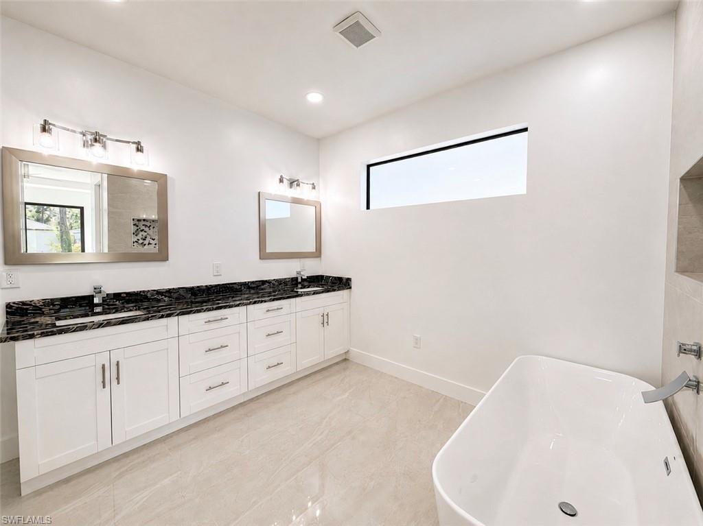Spacious bathroom featuring a freestanding tub, a large vanity with dual sinks, dark stone countertops, white cabinetry, and light tile flooring