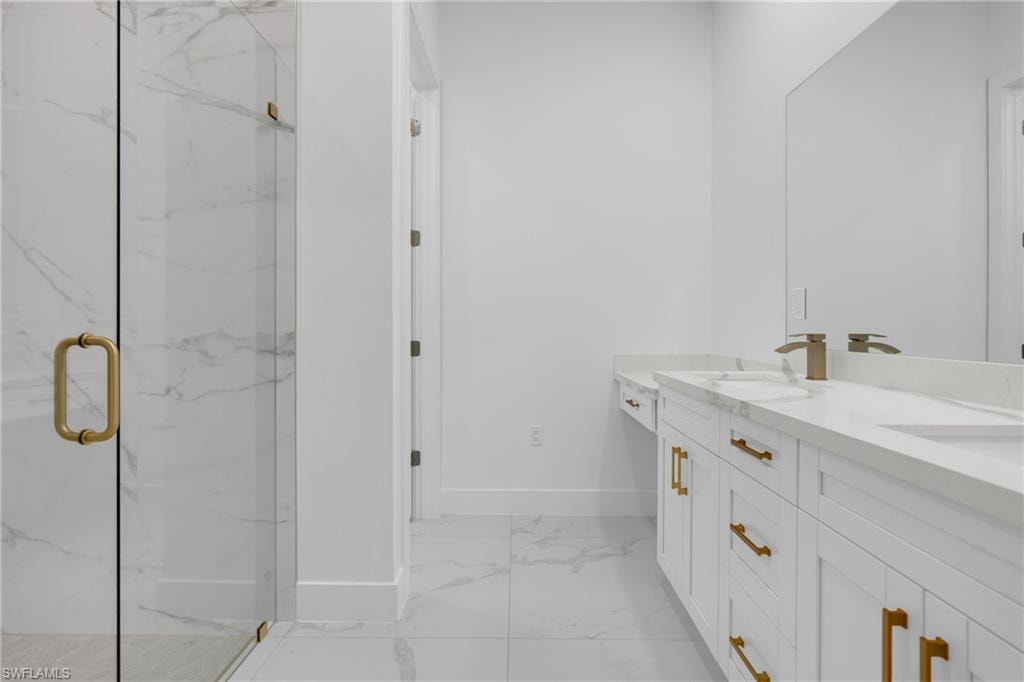 Spacious bathroom featuring a frameless glass shower enclosure, double vanity with integrated sinks, white cabinetry with gold-tone hardware, and large format tile flooring