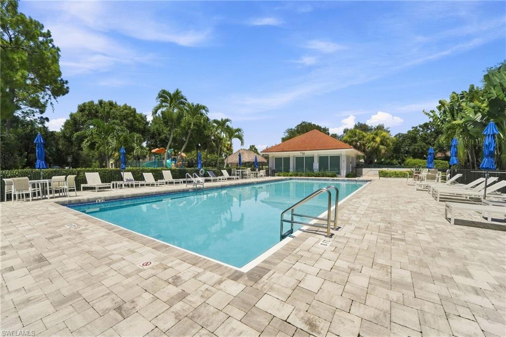 Expansive outdoor pool area with surrounding paver decking, mature tropical landscaping, and a nearby structure featuring a red tile roof