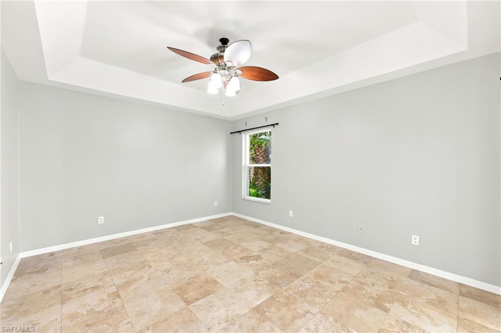 Room with tray ceiling, ceiling fan with integrated lighting, window with exterior foliage, light gray walls, and large format floor tiles