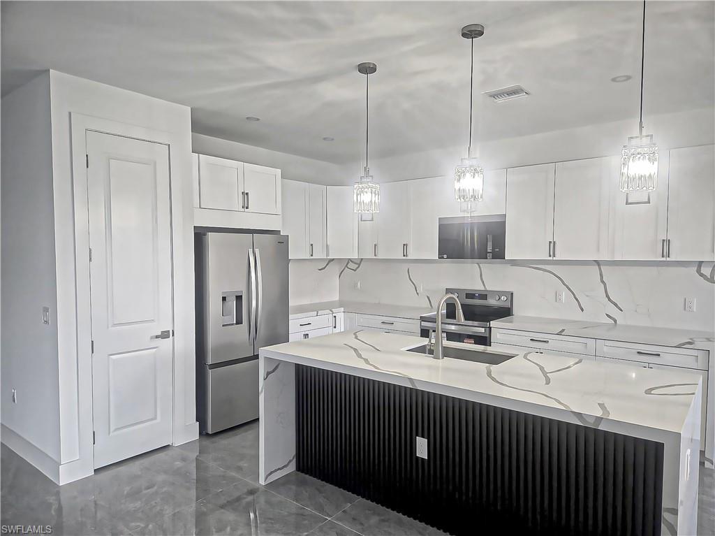 Sleek kitchen featuring white cabinetry, stone countertops, stainless steel appliances, a center island with a built-in sink, and pendant lighting