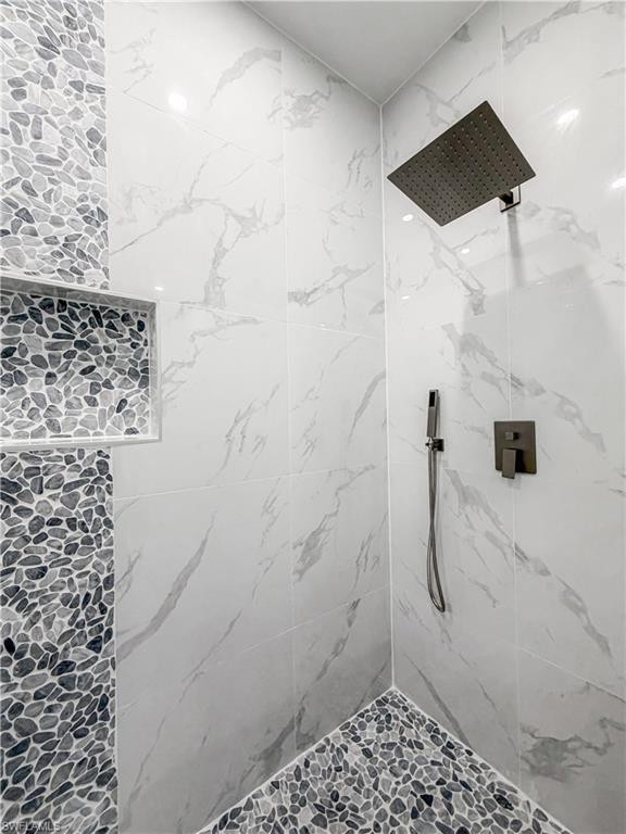Shower enclosure featuring large format white marble-look wall tiles, pebble-tiled accent wall and flooring, square rain shower head, and built-in shower niche
