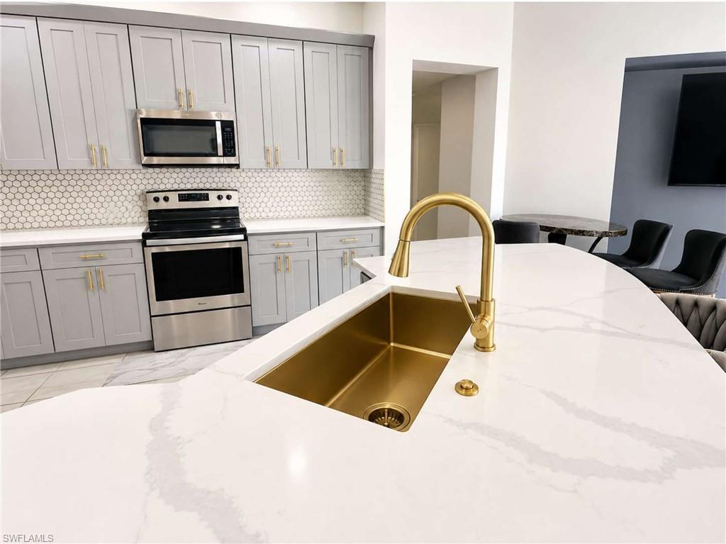 Kitchen featuring light grey cabinetry, a white hexagonal tile backsplash, stainless steel appliances, a white countertop with grey veining, and a gold-finish sink with matching faucet