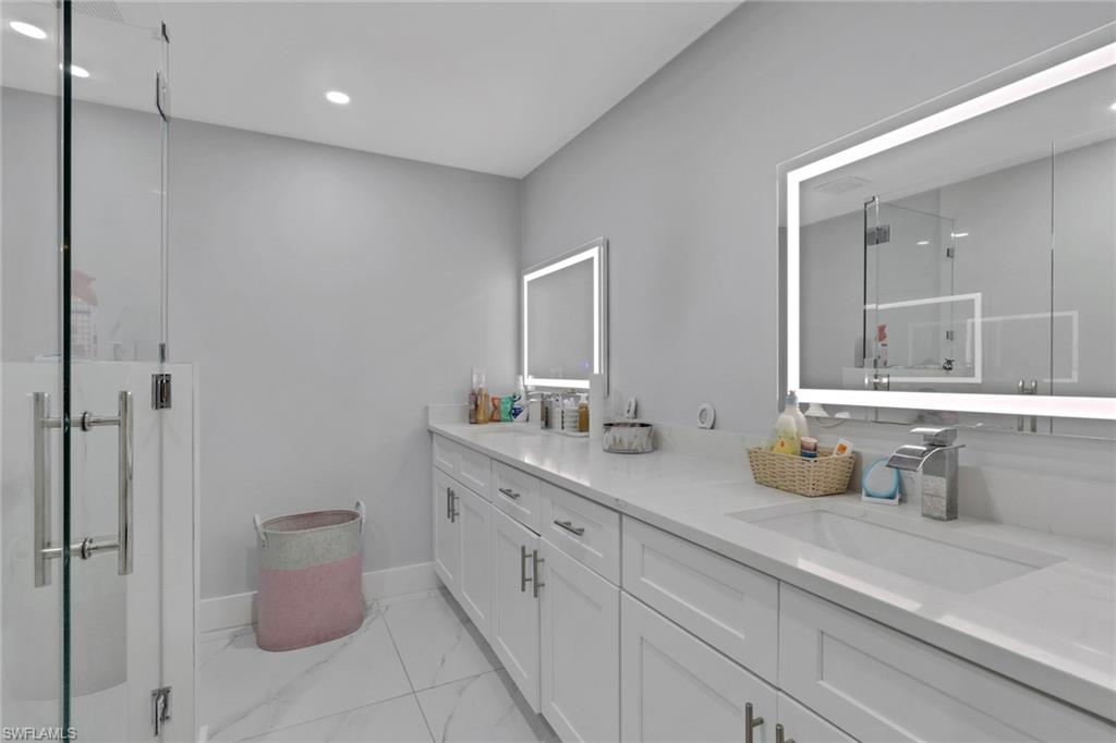 Dual vanity with integrated sinks, white cabinetry, illuminated mirrors, a glass-enclosed shower, and large format floor tiling