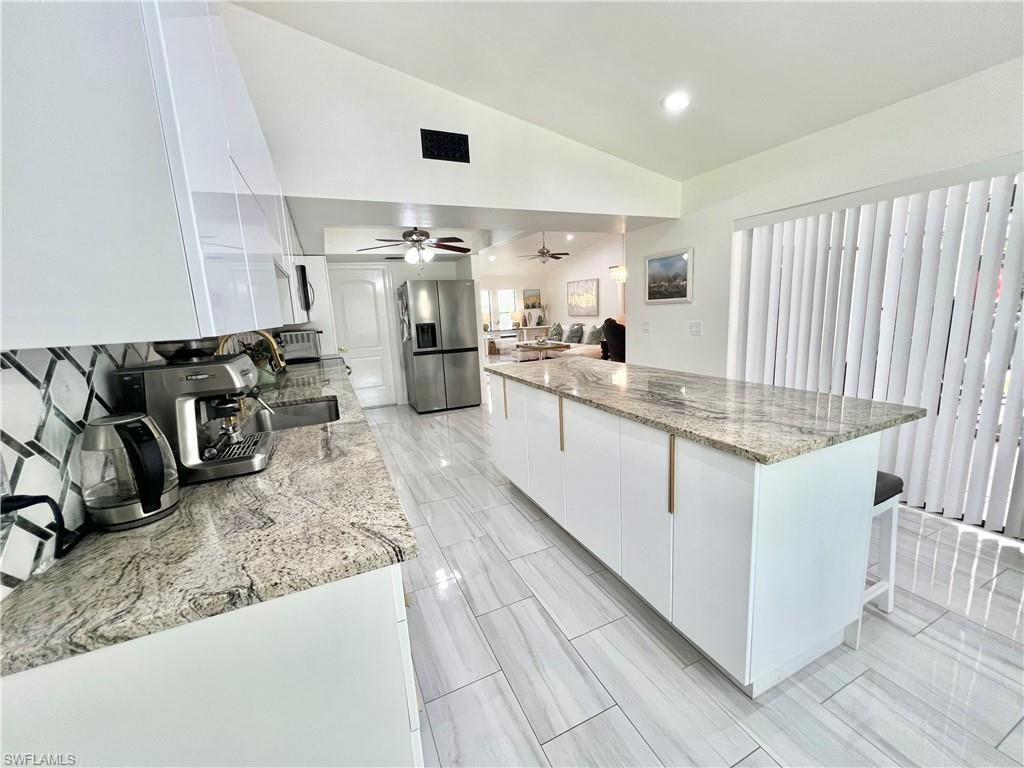 Modern kitchen featuring a spacious island with a light-toned granite countertop, white cabinetry with linear gold-toned pulls, a geometric tile backsplash, and large format tile flooring