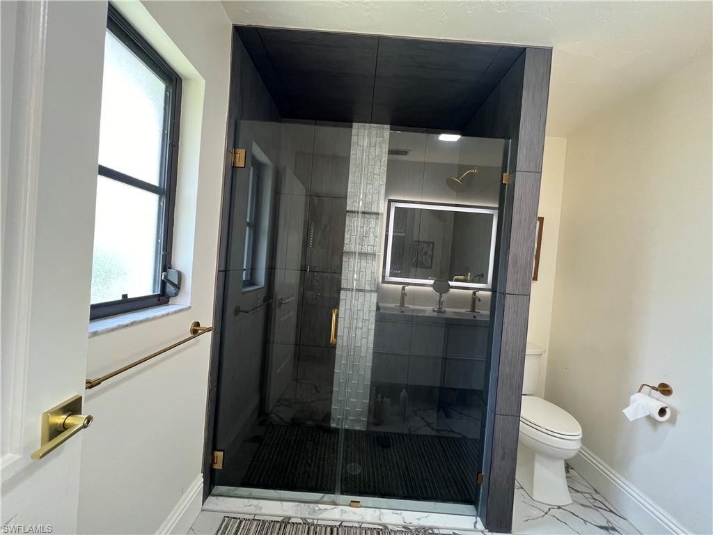Modern bathroom featuring a frameless glass shower enclosure with brass hardware, a dark-finish vanity with dual sinks, an illuminated mirror, and marble-look floor tiling