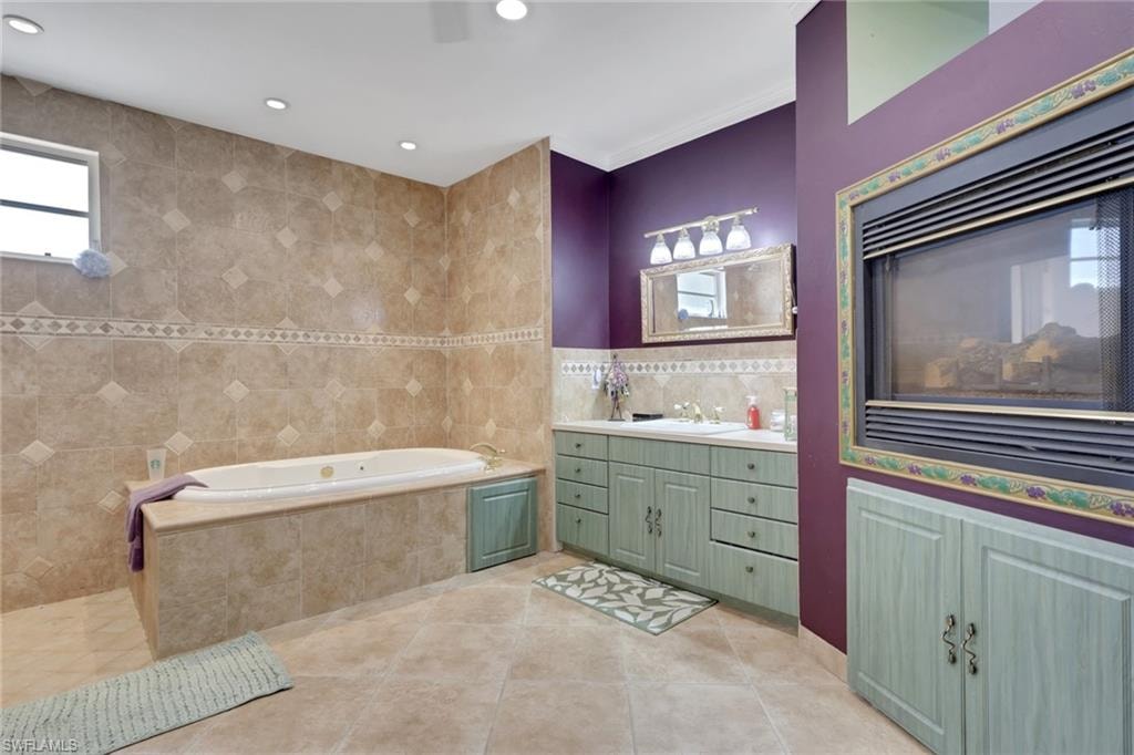 Bathroom featuring a built-in jetted tub, extensive tilework, a double vanity with light green cabinetry, and an integrated fireplace