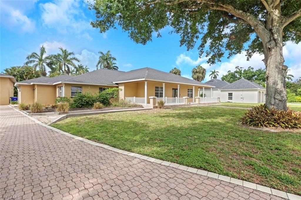 Expansive paver driveway leading to a stucco residence featuring a covered front porch with white railing, a detached structure with a hip roof, and mature landscaping including palm trees