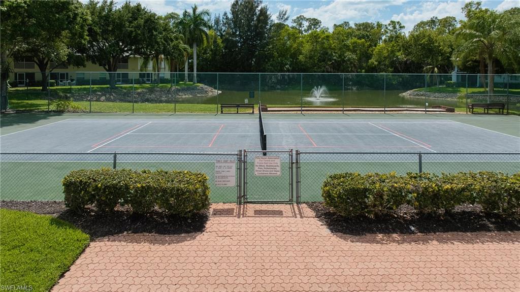 Tennis court with an adjacent paved entryway, chain link fencing, manicured shrubbery, and a water feature with a fountain