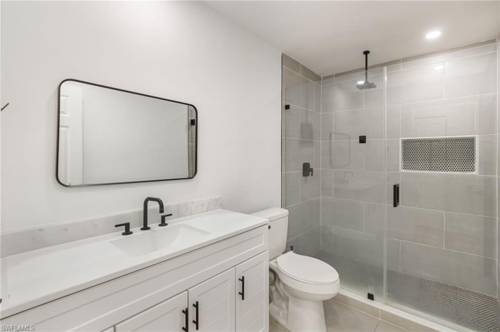 Bathroom featuring a white vanity with a rectangular undermount sink and matte black faucet, a framed rectangular mirror, a toilet, and a glass-enclosed shower with gray tiled walls and a built-in niche