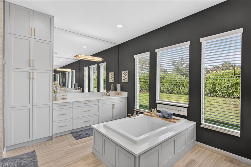 Spacious bathroom featuring a built-in soaking tub, extensive cabinetry, light wood-finish flooring, and multiple windows overlooking a green outdoor space