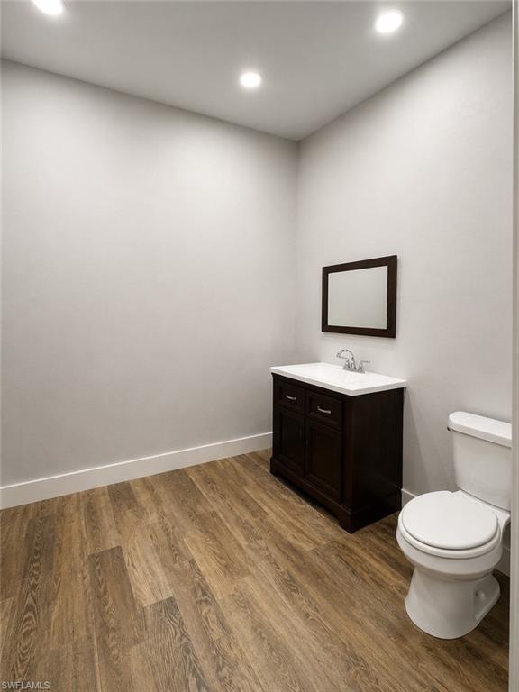 Room with wood-finish flooring, a dark wood vanity with a white countertop and basin sink, a wall-mounted mirror, a white toilet, and recessed lighting