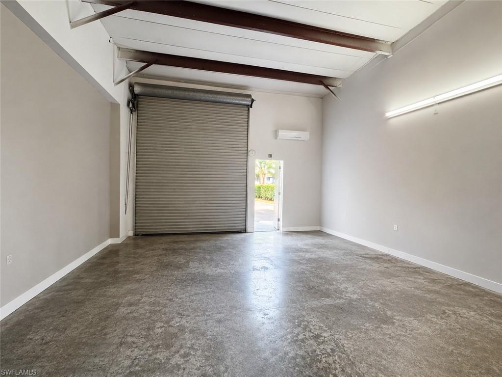 Spacious interior featuring a large roll-up door, exposed ceiling beams, concrete flooring, and a wall-mounted air conditioning unit
