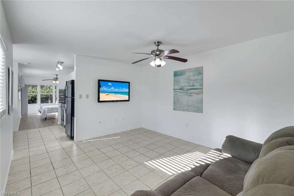 Bright, open living area featuring tile flooring throughout, ceiling fan with integrated lighting, wall-mounted flat-screen television, and abundant natural light from multiple windows