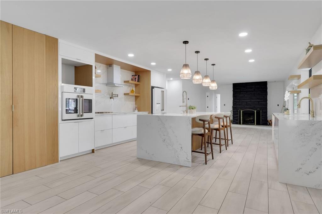 Spacious kitchen featuring an expansive waterfall island, integrated stainless steel appliances, light wood-finish flooring, white cabinetry, and a black brick fireplace with a brass firebox