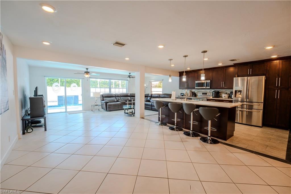 Spacious open-concept interior featuring light-toned tile flooring, a kitchen island with seating, dark wood cabinetry, stainless steel appliances, and sliding glass doors opening to a pool area