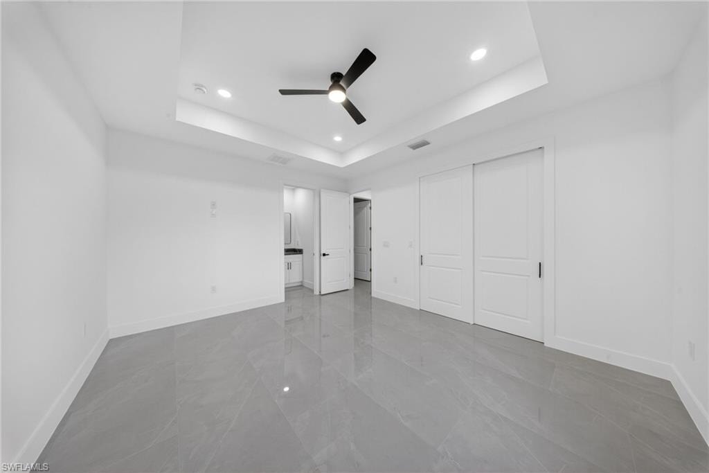 Spacious room featuring large format grey tile flooring, a tray ceiling with recessed lighting, a ceiling fan, and white sliding closet doors