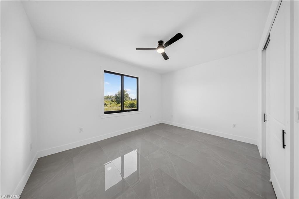 Spacious room featuring a single window with black trim, a ceiling fan with integrated lighting, white walls, and large format grey tile flooring