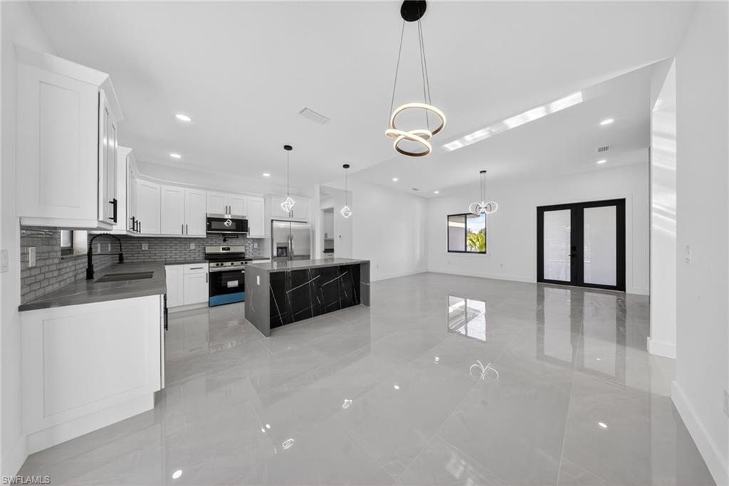 Open-concept kitchen and living area featuring a large central island with a marbled black finish, white shaker cabinetry, stainless steel appliances, and sleek gray tile flooring