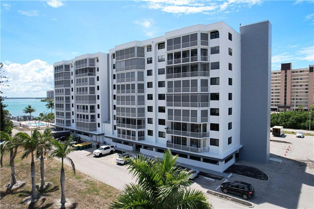 Multi-story condominium building featuring screened balconies, ample surface parking, a waterfront setting, palm trees, and a ground-level swimming pool