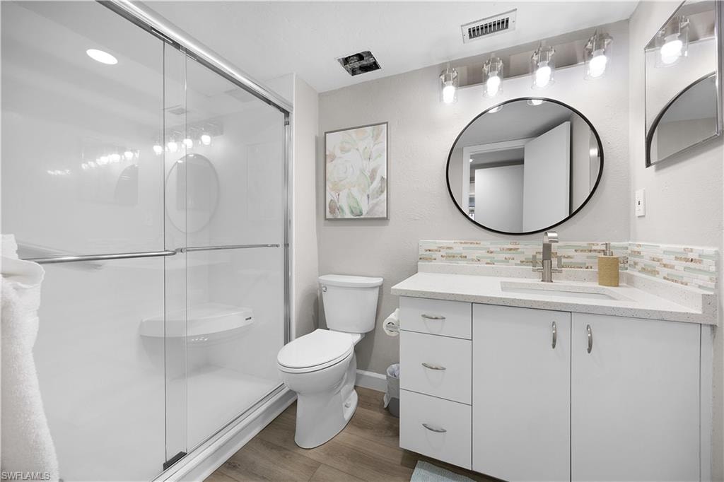 Contemporary bathroom featuring a spacious glass-enclosed shower, wood-finish flooring, a white vanity with silver hardware, a white countertop, and a decorative tile backsplash