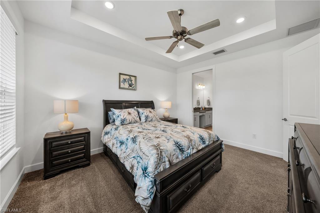 Bedroom featuring a tray ceiling with recessed lighting and a ceiling fan, carpet flooring, a window with blinds, and an entryway to a bathroom with dual vanities