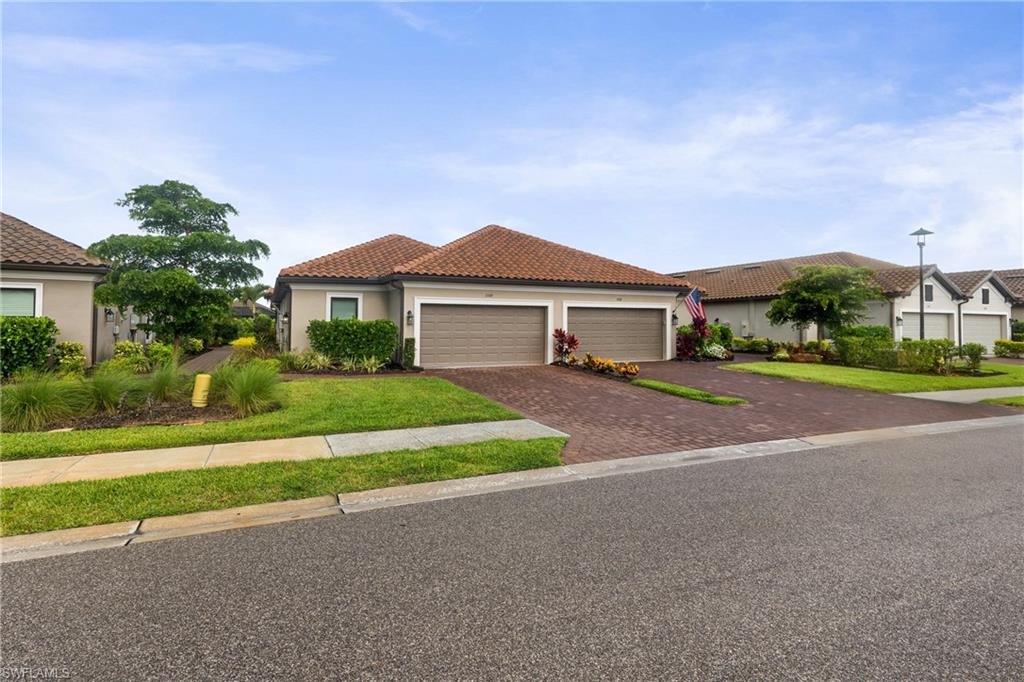 Mediterranean-style architecture featuring a two-car garage with wood-look doors, terracotta roof tiles, stucco exterior, a paver driveway, and landscaped front yard with established plantings