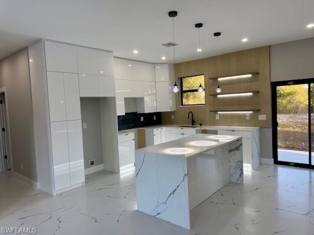 Modern kitchen featuring a waterfall island, sleek white cabinetry, a dark tiled backsplash, wood-finish accent wall with integrated shelving, and recessed lighting