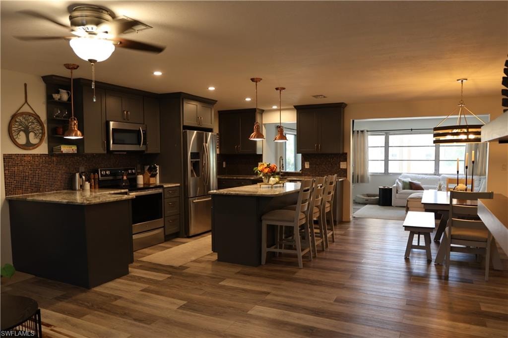 Open-concept kitchen featuring dark cabinetry, stainless steel appliances, granite countertops, and mosaic tile backsplash