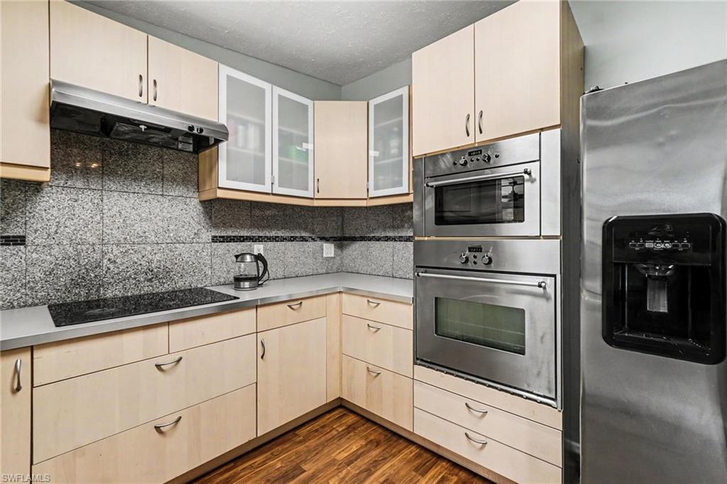 Modern kitchen featuring light wood-finish cabinetry, stainless steel appliances including a built-in oven and microwave, an induction cooktop, and a patterned granite-style backsplash
