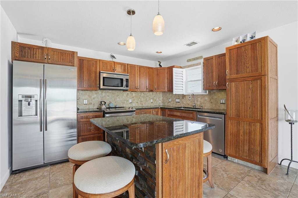 Kitchen featuring wood cabinetry, stainless steel appliances, a stone tile backsplash, a dark countertop island with stone detailing, and recessed lighting