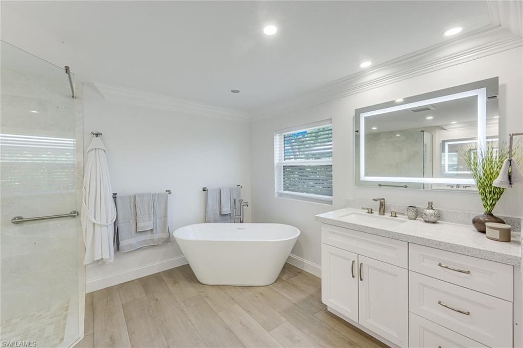 Primary Bathroom featuring a freestanding soaking tub, glass-enclosed shower, wood-finish flooring, a white vanity with a light-toned countertop, and an illuminated mirror