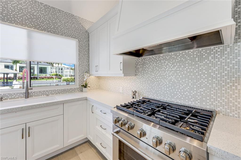 Modern kitchen featuring white shaker cabinetry, light-toned countertops, a full-height tile backsplash with a mosaic pattern, a stainless steel range with a built-in hood, and under-cabinet lighting