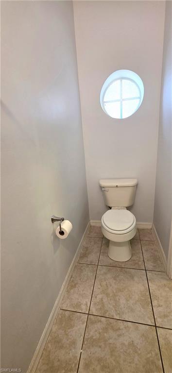 Powder room featuring a circular architectural window, a toilet with a round bowl, light-toned tiled flooring, and a chrome toilet paper holder