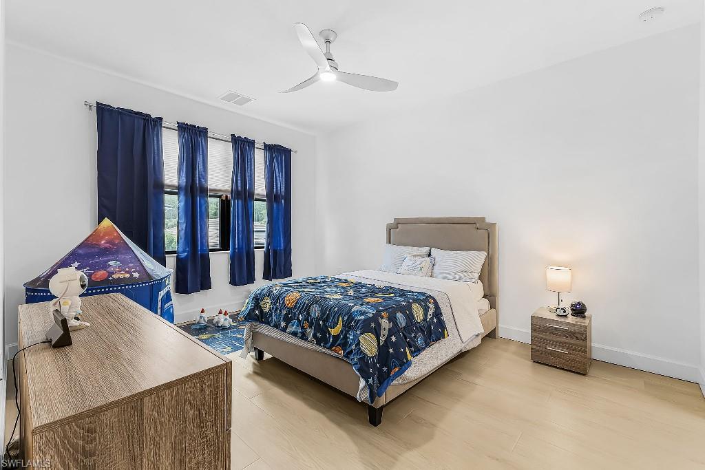 Bright room with light wood-finish flooring, featuring multiple windows with roller shades, a modern ceiling fan, and white baseboards
