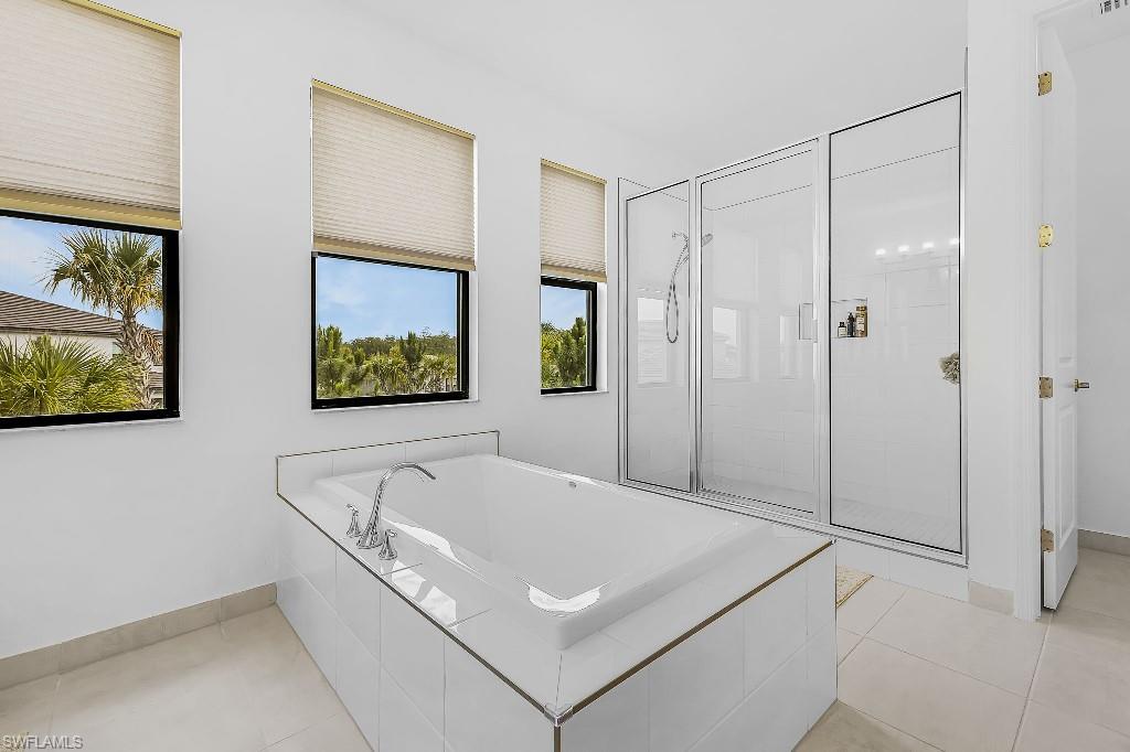 Spacious bathroom featuring a built-in soaking tub with chrome fixtures, a large glass-enclosed shower, multiple windows with blinds, and light-toned tile flooring