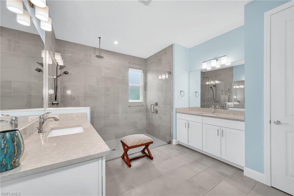 Expansive bathroom featuring dual vanity sinks set in light-toned countertops, a spacious walk-in shower with full-height gray tile, and a rainfall showerhead