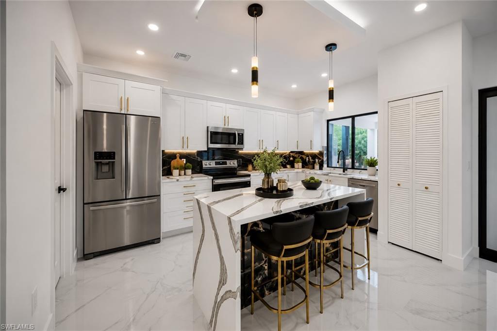 Gourmet kitchen featuring a waterfall island with integrated seating, stainless steel appliances, white cabinetry with gold-tone hardware, a black and white patterned backsplash, and recessed lighting