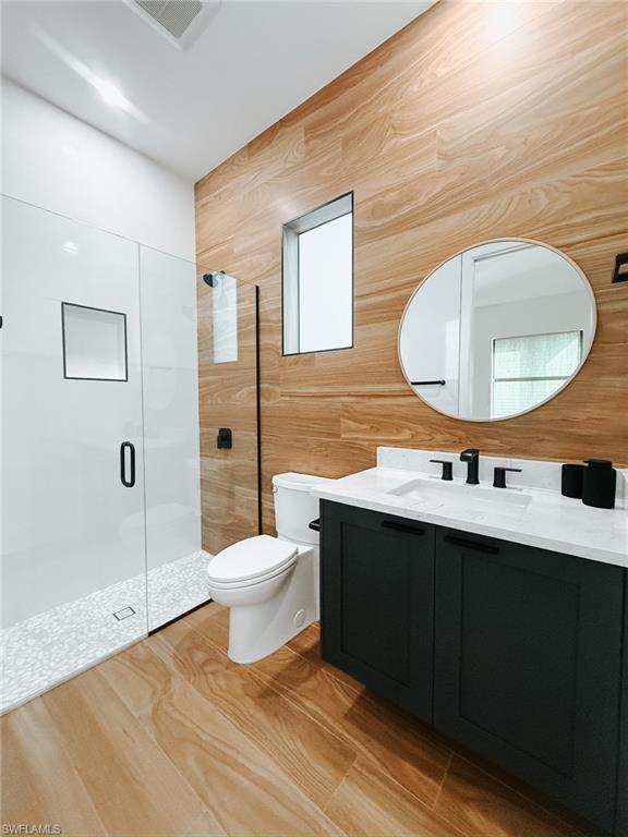 Bathroom featuring wood-finish wall paneling, a dark wood-finish vanity with a white countertop and rectangular sink, matte black fixtures, a frameless oval mirror, and a glass-enclosed shower with pebble-style flooring
