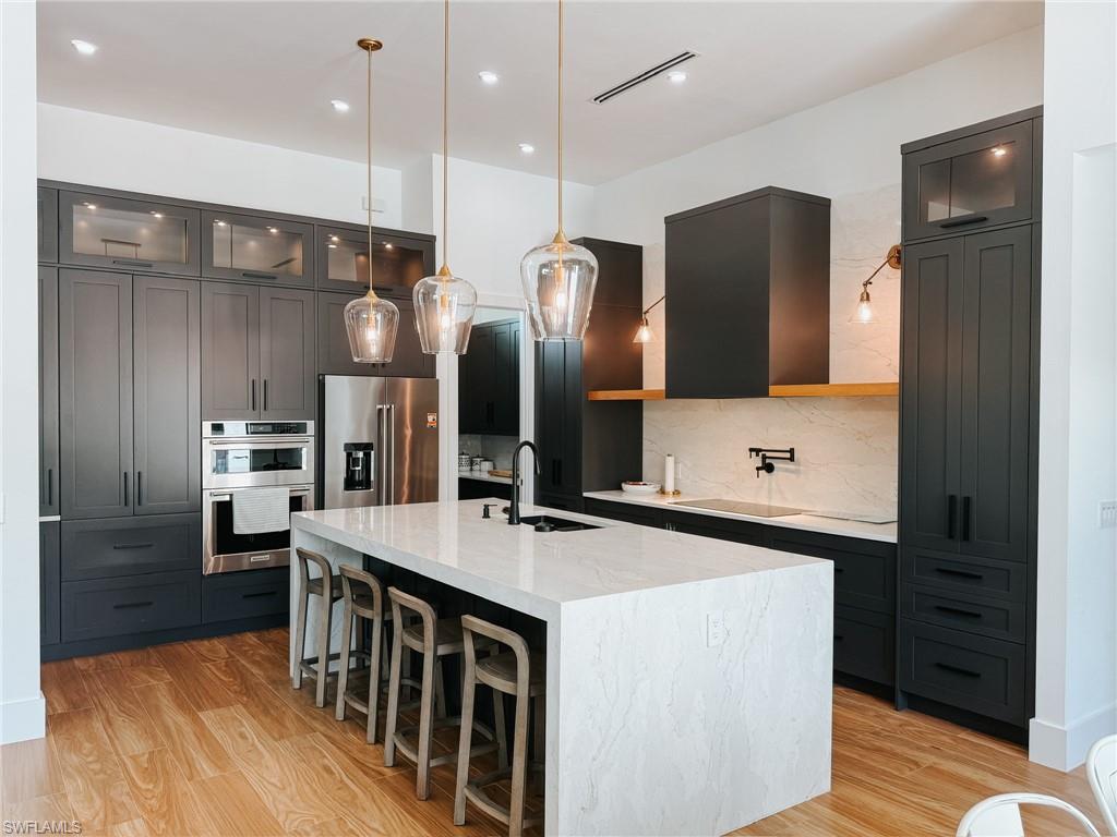 Gourmet kitchen featuring a large waterfall island with integrated sink, dark cabinetry with glass-front uppers, stainless steel appliances, wood-finish flooring, and a white tile backsplash
