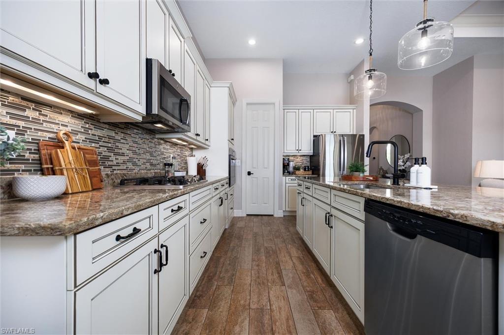 Gourmet kitchen featuring wood-finish flooring, white cabinetry with dark hardware, a mosaic tile backsplash, stainless steel appliances, and a spacious island with a built-in sink and pendant lighting