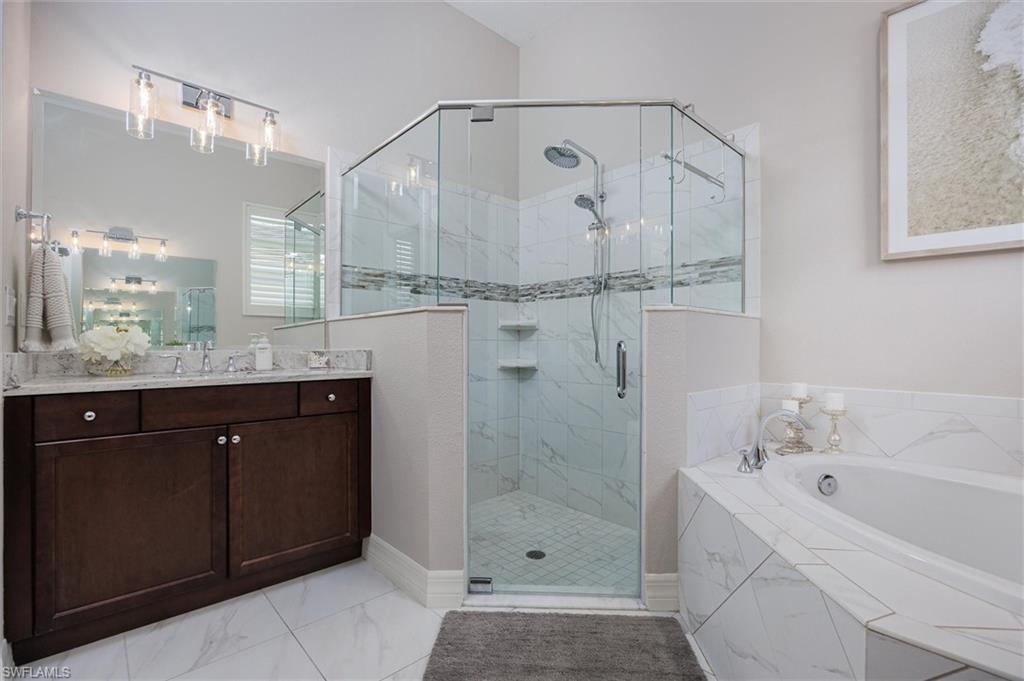 Bathroom featuring a corner soaking tub with a tiled surround, a glass-enclosed shower with tiled walls and built-in shelving, a dual-sink vanity with a stone countertop, and large format tile flooring