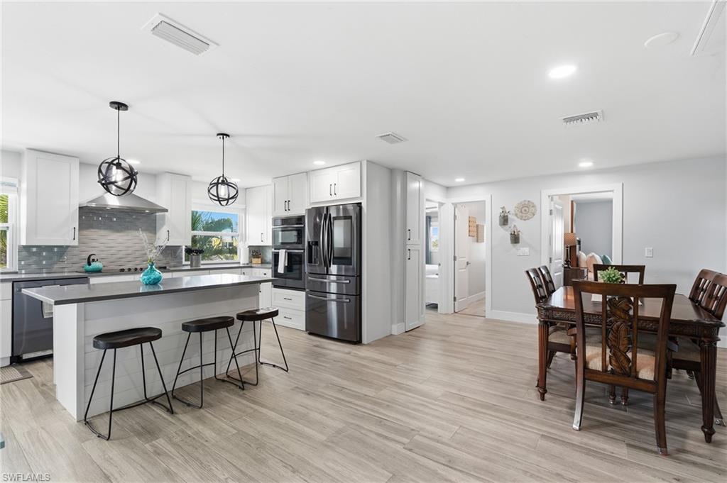 Open-concept living space featuring a kitchen island with seating, stainless steel appliances, white cabinetry, and wood-finish flooring - Virtually Edited Image