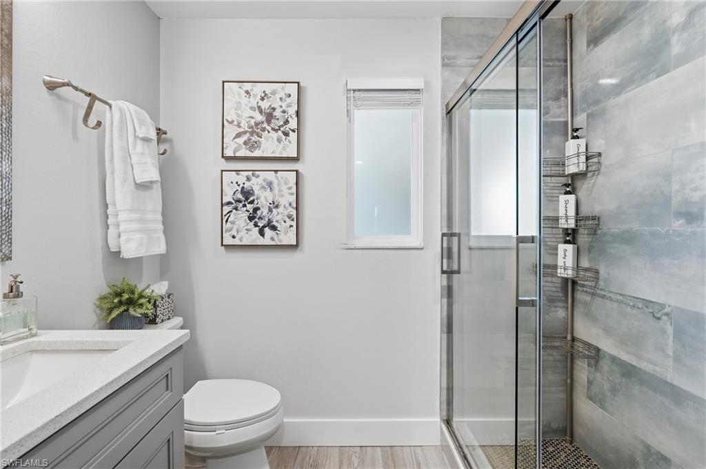 Contemporary bathroom featuring a glass-enclosed shower with tile surround, a single vanity with a solid surface countertop, wood-finish flooring, and a window providing natural light - Virtually Edited Image
