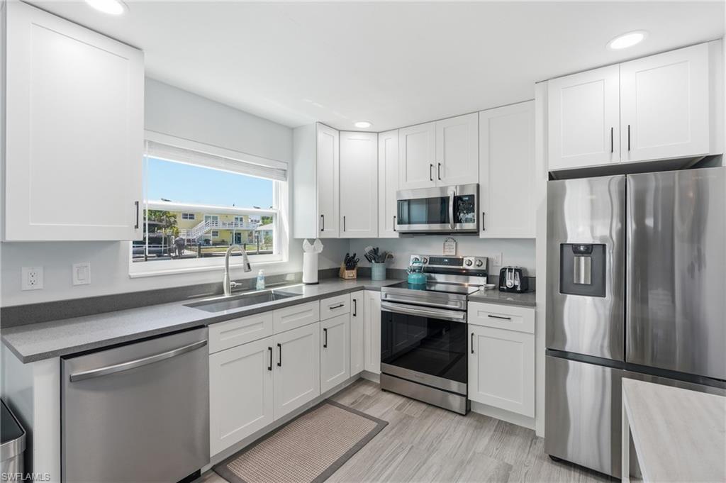 Modern kitchen featuring white shaker cabinetry, stainless steel appliances, gray countertops, a stainless steel sink with a gooseneck faucet, and light wood-finish flooring - Virtually Edited Image