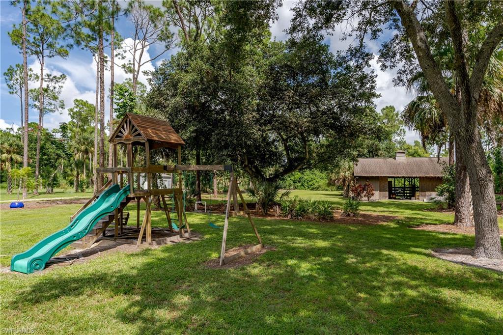 Expansive outdoor space featuring a wooden play structure with a slide and swings, mature trees, a manicured lawn, and a distant covered pavilion with a pitched roof