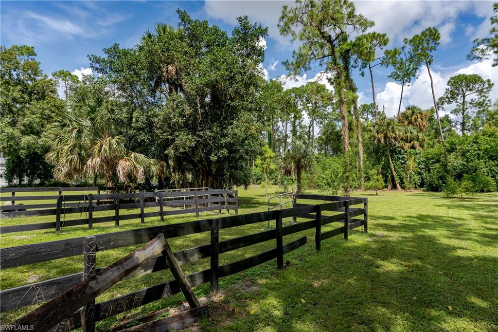Expansive fenced pasture featuring a dark wood rail fence, lush green grass, a variety of mature trees, and a partially cloudy sky