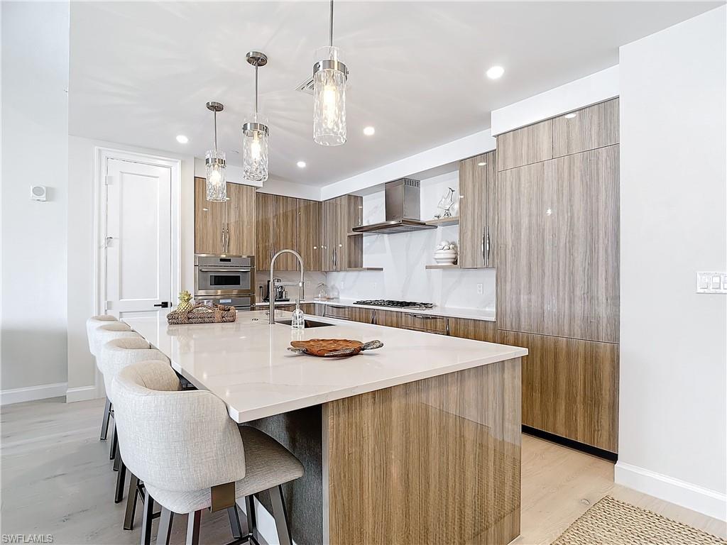 Kitchen with appliances with stainless steel finishes, a large island, light hardwood / wood-style floors, wall chimney exhaust hood, and pendant lighting