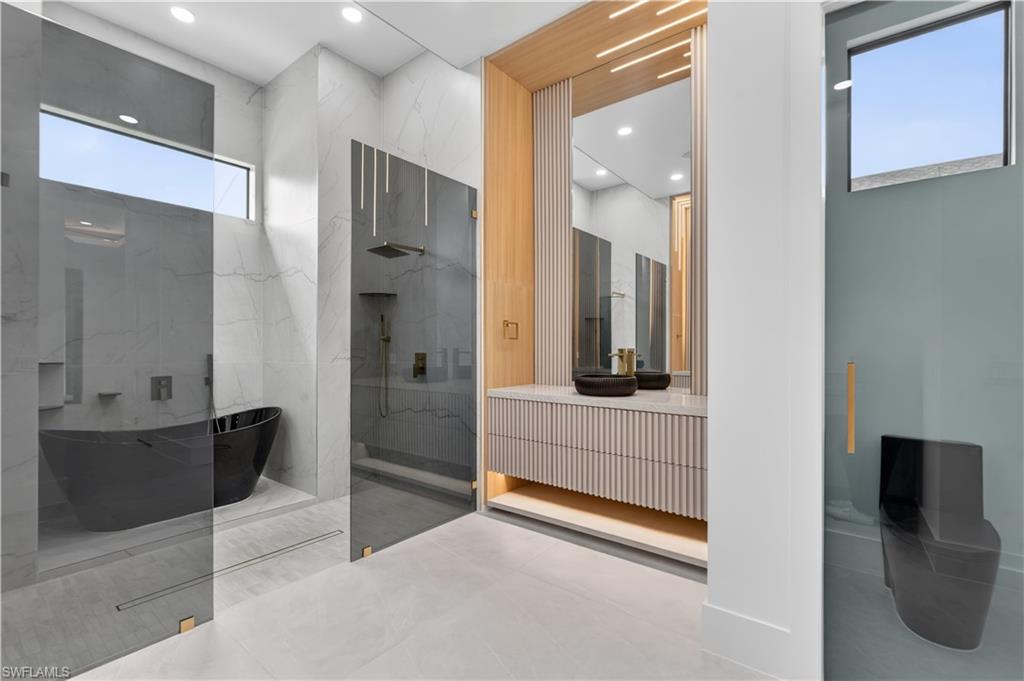 Modern bathroom featuring a freestanding black soaking tub, a frameless glass shower enclosure with a rain shower head, a floating vanity with reeded paneling and integrated lighting, and a private toilet area